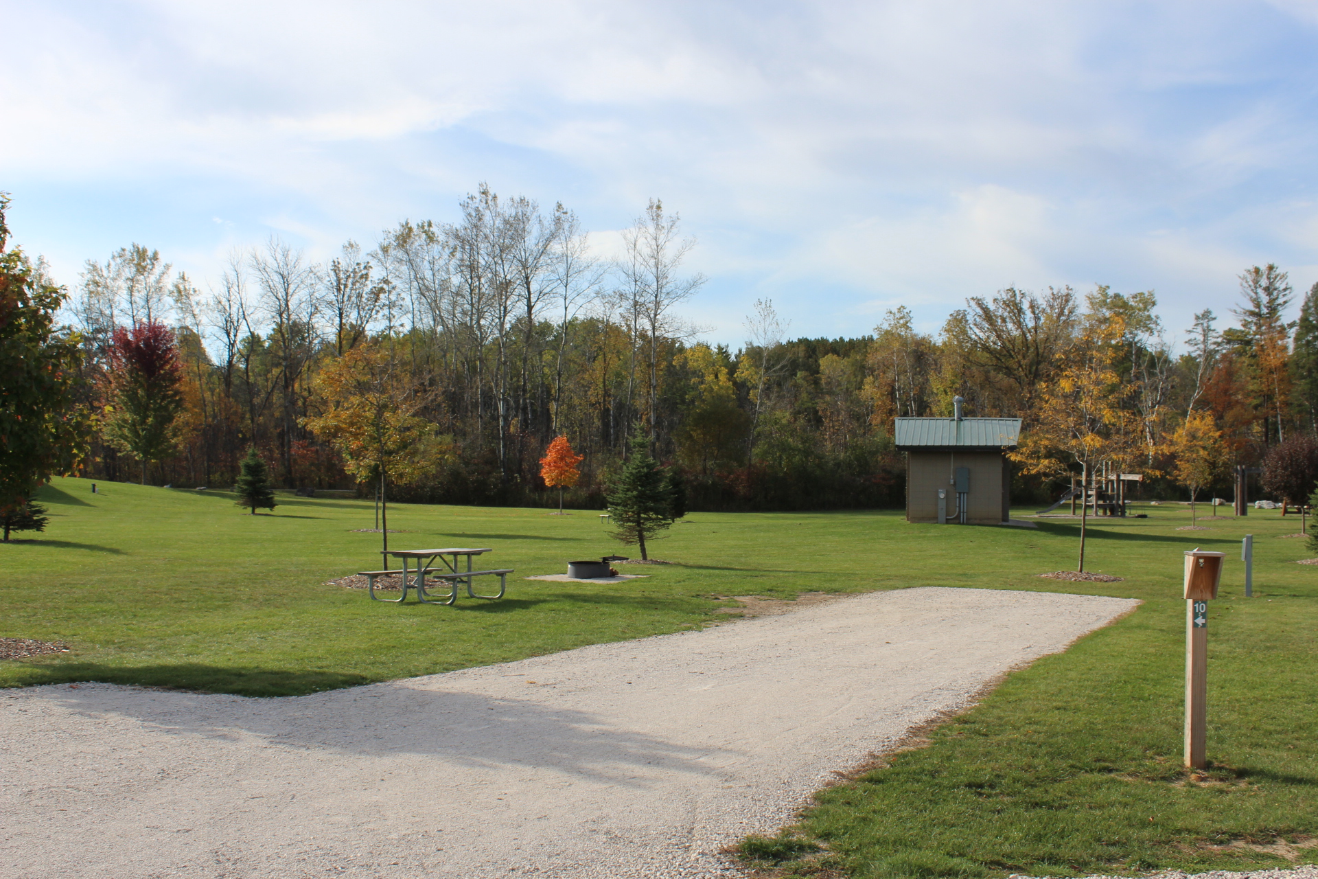 A grassy outdoor campground site with a gravel pad, a picnic table on the lawn next to a fire ring, and mature trees lining the background.