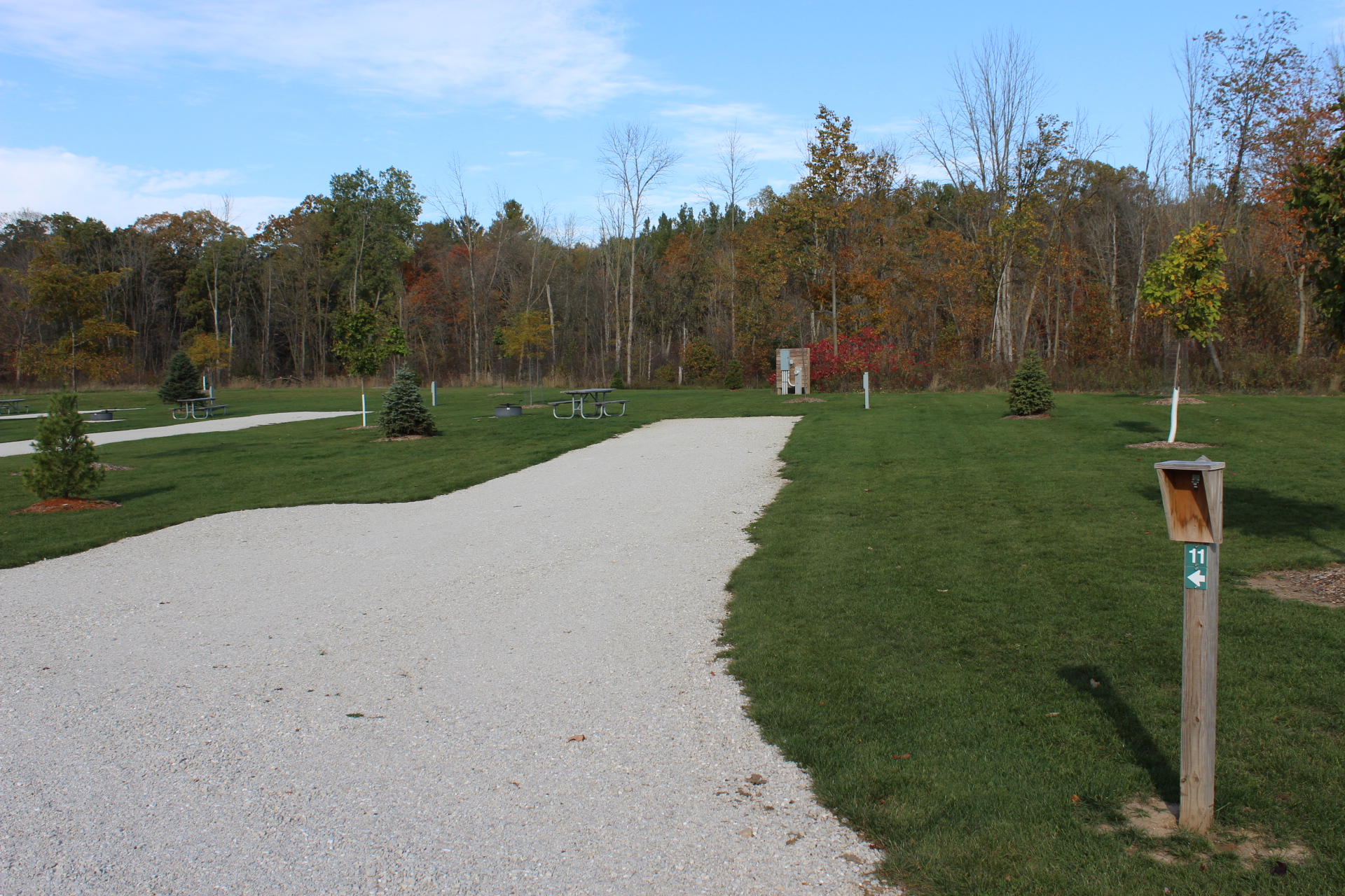 A grassy outdoor campground site with a gravel pad, a picnic table on the lawn next to a fire ring, and mature trees lining the background.