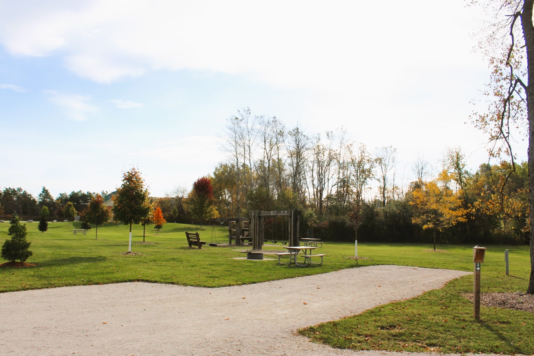 A grassy outdoor campground site with a gravel pad, a picnic table on the lawn next to a fire ring, and mature trees lining the background.