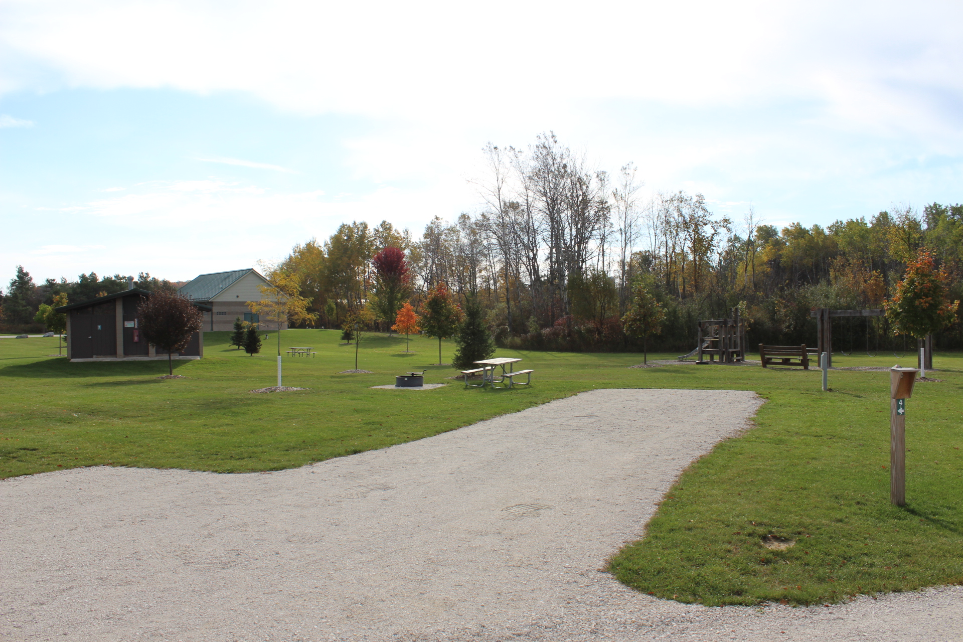 A grassy outdoor campground site with a gravel pad, a picnic table on the lawn next to a fire ring, and mature trees lining the background.
