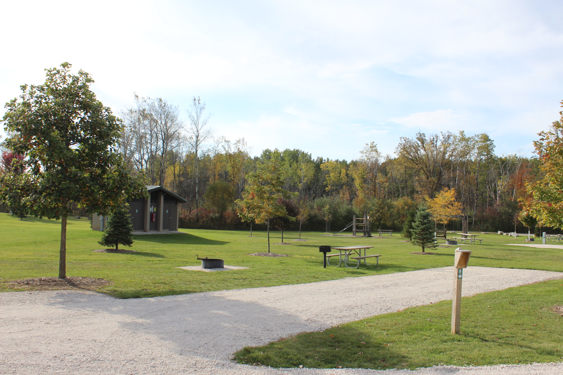 A grassy outdoor campground site with a gravel pad, a picnic table on the lawn next to a fire ring, and mature trees lining the background.